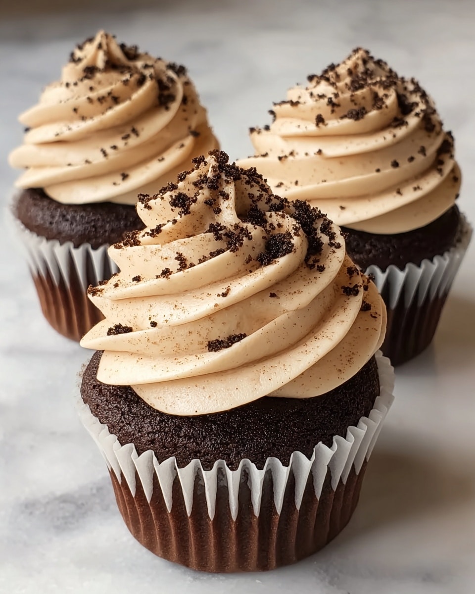 Three chocolate cupcakes sit on a white marbled surface, each wrapped in a white paper liner. Each cupcake has one thick layer of dark brown, soft, moist cake topped with a tall, swirled layer of light beige, creamy frosting. The frosting is sprinkled with small, dark chocolate crumb bits scattered mainly on top and slightly down the sides. The cupcakes are close to each other with one in front and two slightly behind. Photo taken with an iphone --ar 4:5 --v 7