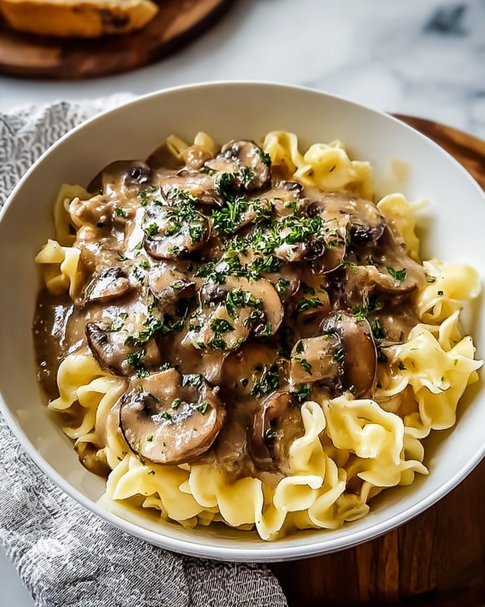 A white bowl holds a creamy pasta dish with two main layers. The bottom layer is soft, light yellow pasta pieces with a slightly wavy texture. On top, there is a thick mushroom sauce with brown sliced mushrooms covered in a shiny, smooth gravy. The dish is finished with finely chopped green herbs sprinkled over the mushrooms. The bowl is set on a white marbled surface with a light gray patterned cloth nearby. photo taken with an iphone --ar 4:5 --v 7