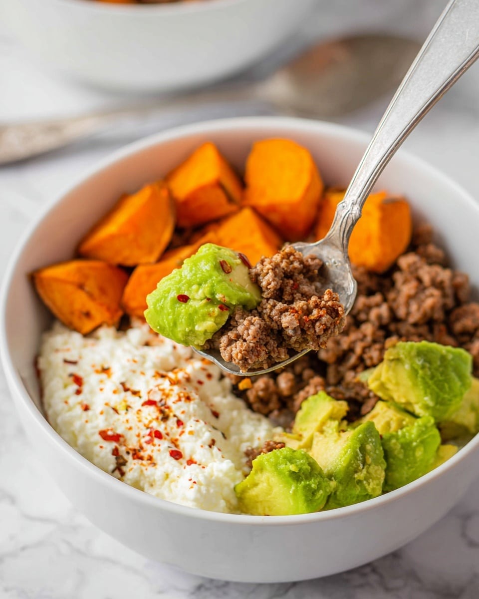 A white bowl holds a dish with four main layers: bright green chunks of avocado on the right side, crumbly brown cooked ground beef below the avocado, vibrant orange roasted sweet potato cubes on the left, and a creamy white cottage cheese with a sprinkling of red chili flakes next to the sweet potatoes. A silver spoon lifts a mix of all four ingredients toward the camera, showing a close-up of the green avocado, orange sweet potato, brown beef, and white cottage cheese together. The bowl and spoon rest on a white marbled texture. photo taken with an iphone --ar 4:5 --v 7