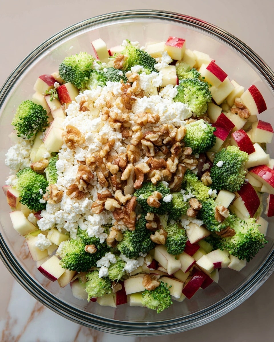 The image shows a clear glass bowl filled with a colorful, mixed salad. The bottom layer is white, crumbly cottage cheese spread evenly. On top, there are small, bright green broccoli florets scattered all over. Next, diced pieces of red and white apple chunks are mixed in evenly. There are also many pieces of chopped walnuts sprinkled across the salad, adding a rough, textured look. The bowl sits on a white marbled surface. The photo taken with an iphone --ar 4:5 --v 7