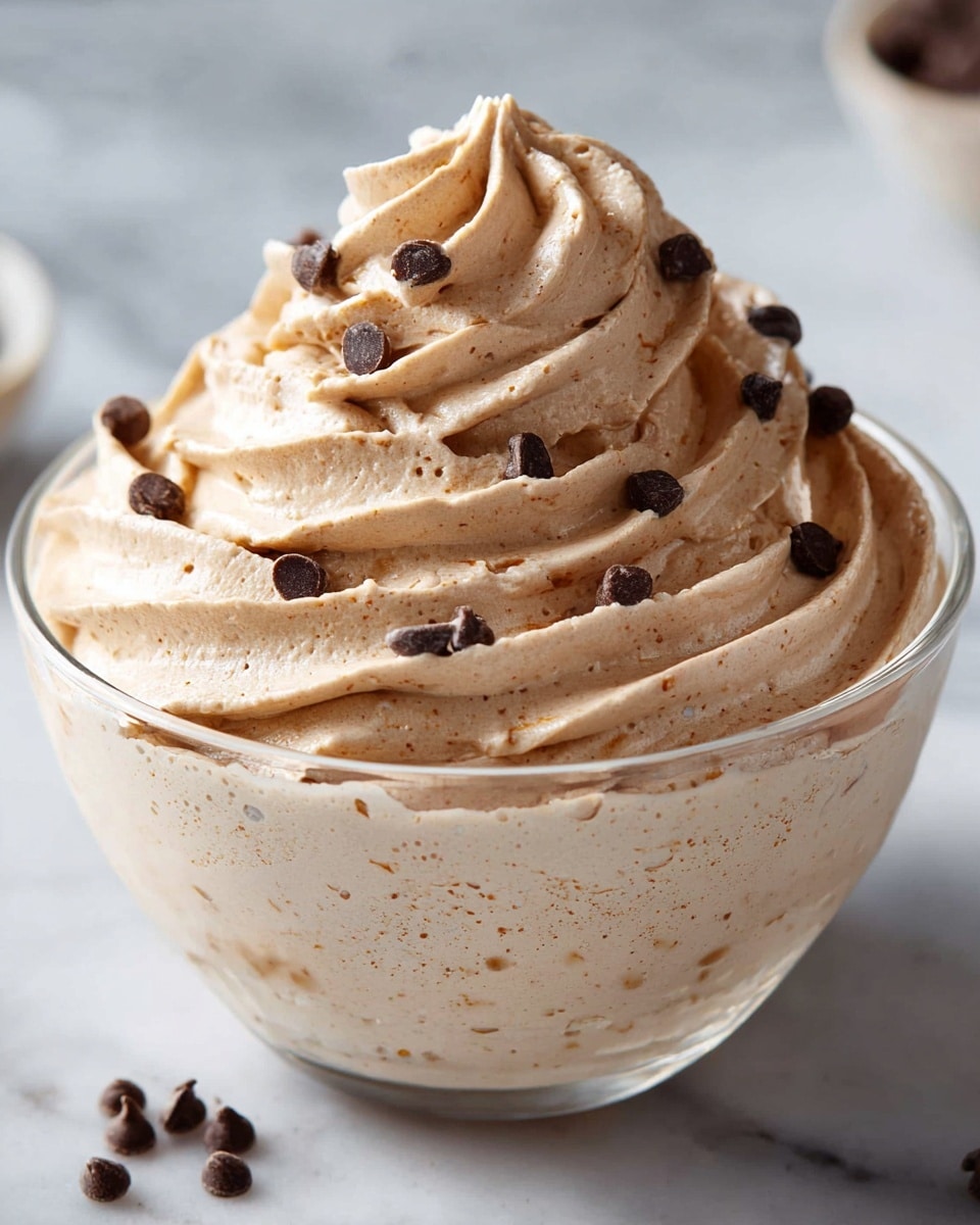 A clear glass bowl filled with a thick, creamy light brown mousse that has a smooth, slightly swirled texture visible on the sides. On top, there is a high, decorative swirl of the same mousse, piped in a circular motion with ridges and a pointed peak at the center. Scattered over the mousse are small, dark brown chocolate chips adding contrast to the light brown cream. The bowl is placed on a white marbled surface, creating a clean and elegant look. Photo taken with an iphone --ar 4:5 --v 7