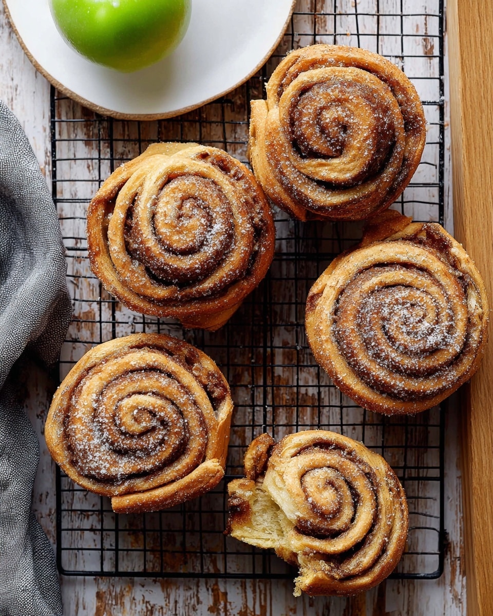 Five cinnamon rolls sit on a black wire cooling rack over a wooden table with a white marbled texture. Each roll is tightly coiled with visible layers of golden-brown dough and dark cinnamon filling, sprinkled evenly with a light dusting of white sugar. The rolls have a soft, slightly crispy texture on the outside, showing the swirled pattern clearly. A green apple is placed on a white plate in the top left corner, and a gray cloth is partially visible in the bottom left. One roll is torn open on the right edge, showing the inner soft and fluffy layers. Photo taken with an iphone --ar 4:5 --v 7
