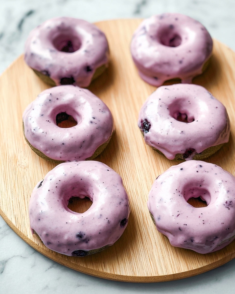Six donuts are placed on a light wooden round board with soft wood grain. Each donut has one visible layer of soft, smooth, pale purple icing with small darker purple specks that slightly drips over the edges. The donut base looks dense and dark green with some visible spots of dark berries inside. The background is a white marbled texture. photo taken with an iphone --ar 4:5 --v 7
