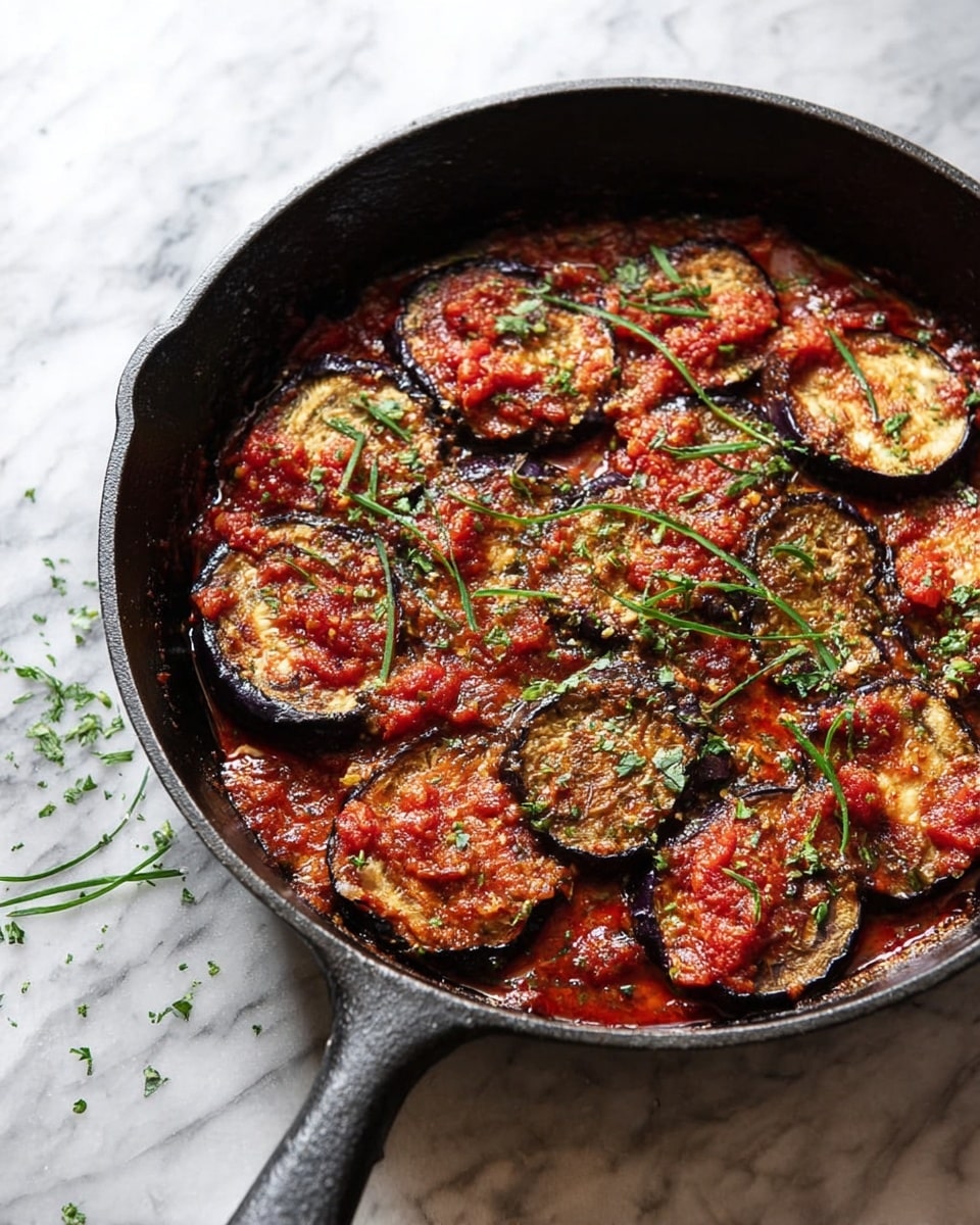 A black cast iron skillet filled with a cooked dish featuring a single layer of round, browned eggplant slices with dark edges, each topped with a chunky red tomato sauce. The eggplant slices are arranged flat and closely packed, showing a mix of light brown, tan, and purple shades. Fresh green herb strips are scattered over the top, adding a touch of color contrast against the rich red sauce and dark eggplant. The skillet sits on a white marbled surface with a few small herb pieces nearby. Photo taken with an iphone --ar 4:5 --v 7
