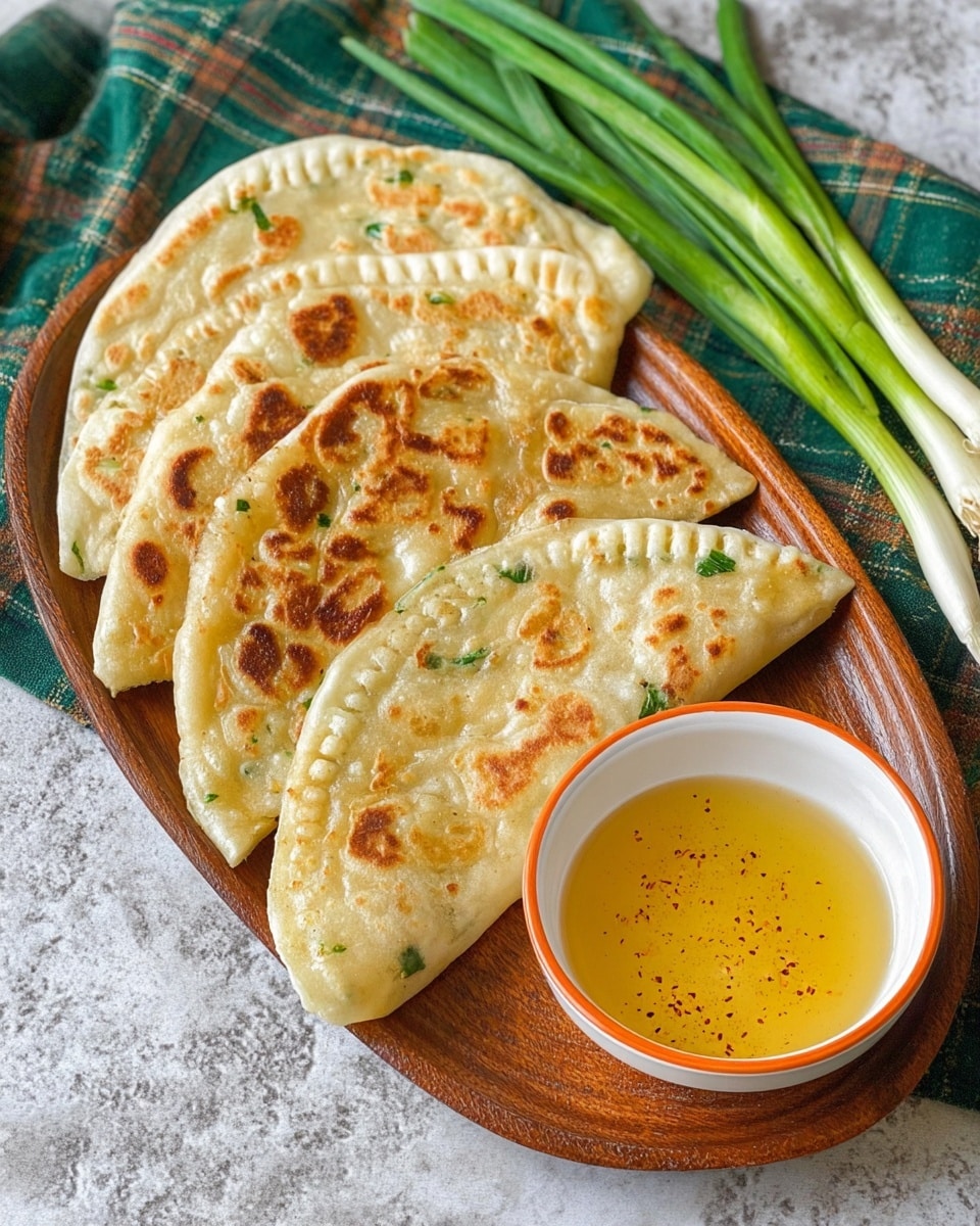 The image shows three layered flatbreads with golden brown spots on their light beige surface, arranged diagonally on a wooden oval plate. The bottom layer is the largest with a consistent thin crust and crimped edges, the middle layer is medium-sized with more pronounced browning and crimping, and the top layer is the smallest and has a slightly folded appearance with green herbs visible inside. On the right side of the plate is a white bowl with an orange rim filled with light yellow dipping sauce speckled with tiny spices. In the background, green onions lay on a green and beige plaid cloth over a white marbled surface. Photo taken with an iphone --ar 4:5 --v 7