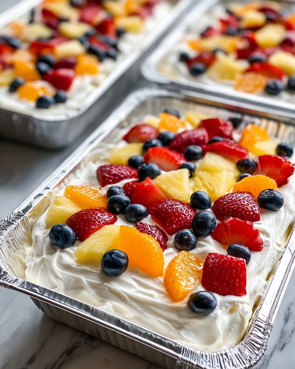 Three foil trays filled with a layered dessert set on a white marbled surface, each tray containing three visible layers: a bottom layer that is not visible but implied, a middle layer of smooth white whipped cream with soft peaks, and a top layer of assorted fresh fruits. The fruit layer features evenly spaced, vibrant red strawberry halves, bright orange mandarin slices, large pale yellow pineapple chunks, and small round dark blue blueberries spread across the whipped cream. The focus is on the front tray, capturing the glossy texture and fresh appearance of the fruits with the shiny foil edges reflecting light around the dessert. Photo taken with an iphone --ar 4:5 --v 7
