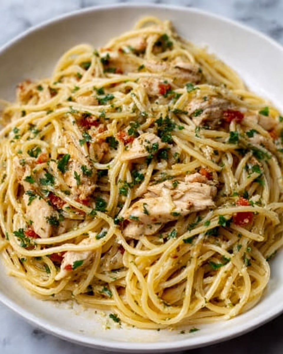 A white plate filled with a serving of spaghetti pasta mixed with small pieces of cooked chicken. The pasta is light yellow with a slight shine, showing a moist texture, and is spread evenly across the plate. Scattered on top are finely chopped green herbs and small bits of red tomato, adding color contrast. The dish looks warm and freshly served, sitting on a white marbled surface. photo taken with an iphone --ar 4:5 --v 7