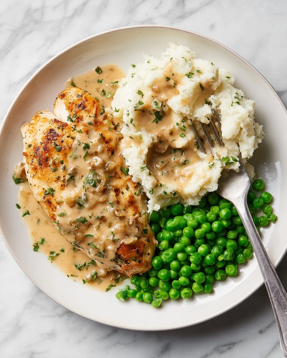 A white plate holds a meal with three main layers: on the left, a piece of golden-brown cooked chicken breast covered with a creamy beige gravy speckled with small bits of onion and herbs; in the top-right section, a fluffy mound of white mashed potatoes topped and partially covered with the same creamy gravy; in the bottom-right section, a pile of bright green peas with a smooth, shiny texture. A silver fork is placed on the right edge of the plate, resting partly on the mashed potatoes. The plate sits on a white marbled surface. Photo taken with an iphone --ar 4:5 --v 7