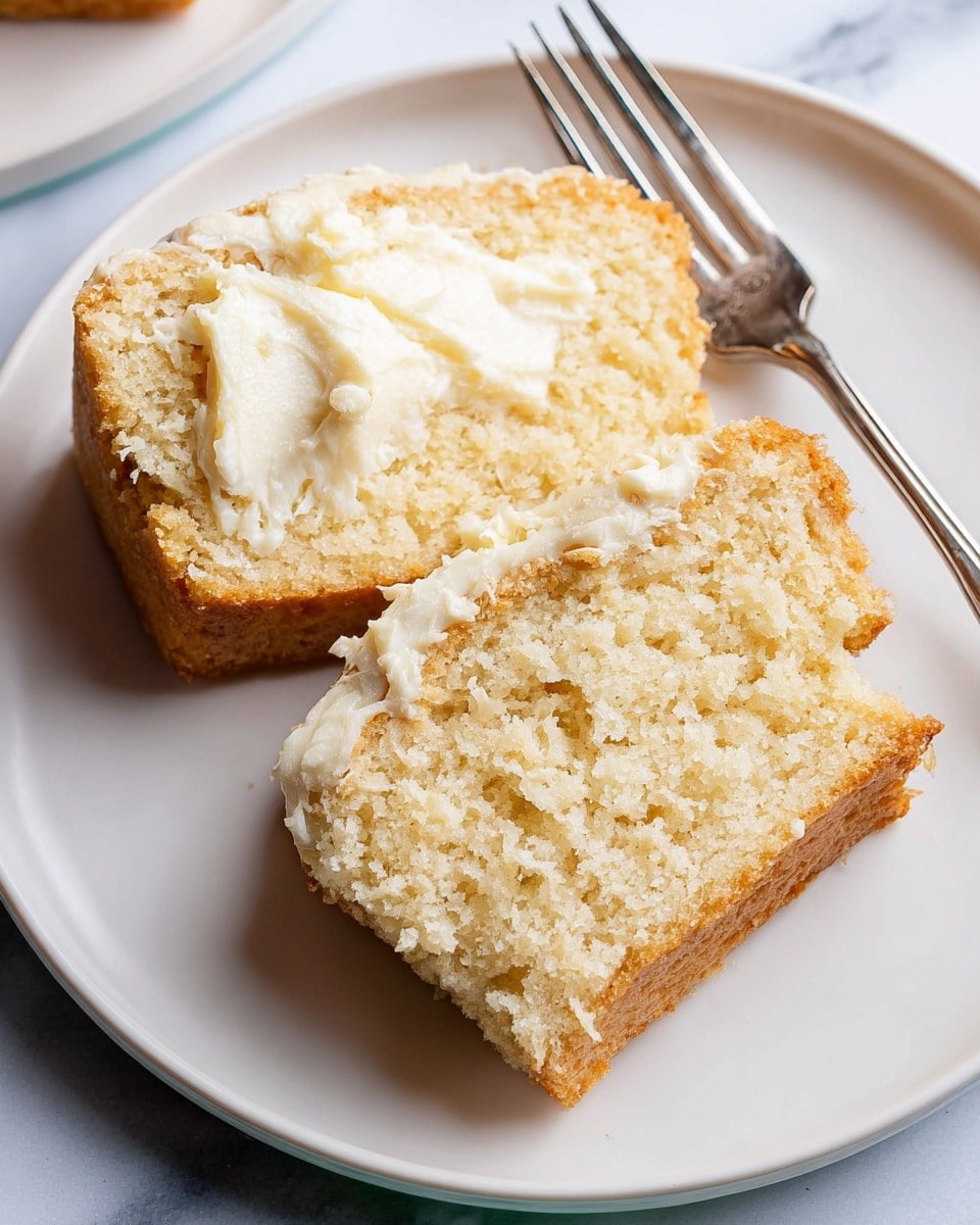 Two slices of light golden brown cake are placed on a white plate with a silver fork nearby. The top slice is spread with a creamy, soft white butter layer in the middle, showing a soft and moist texture inside the cake. The cake crumb looks tender and slightly crumbly, with a smooth golden crust on top and edges. The setting is on a white marbled surface. photo taken with an iphone --ar 4:5 --v 7