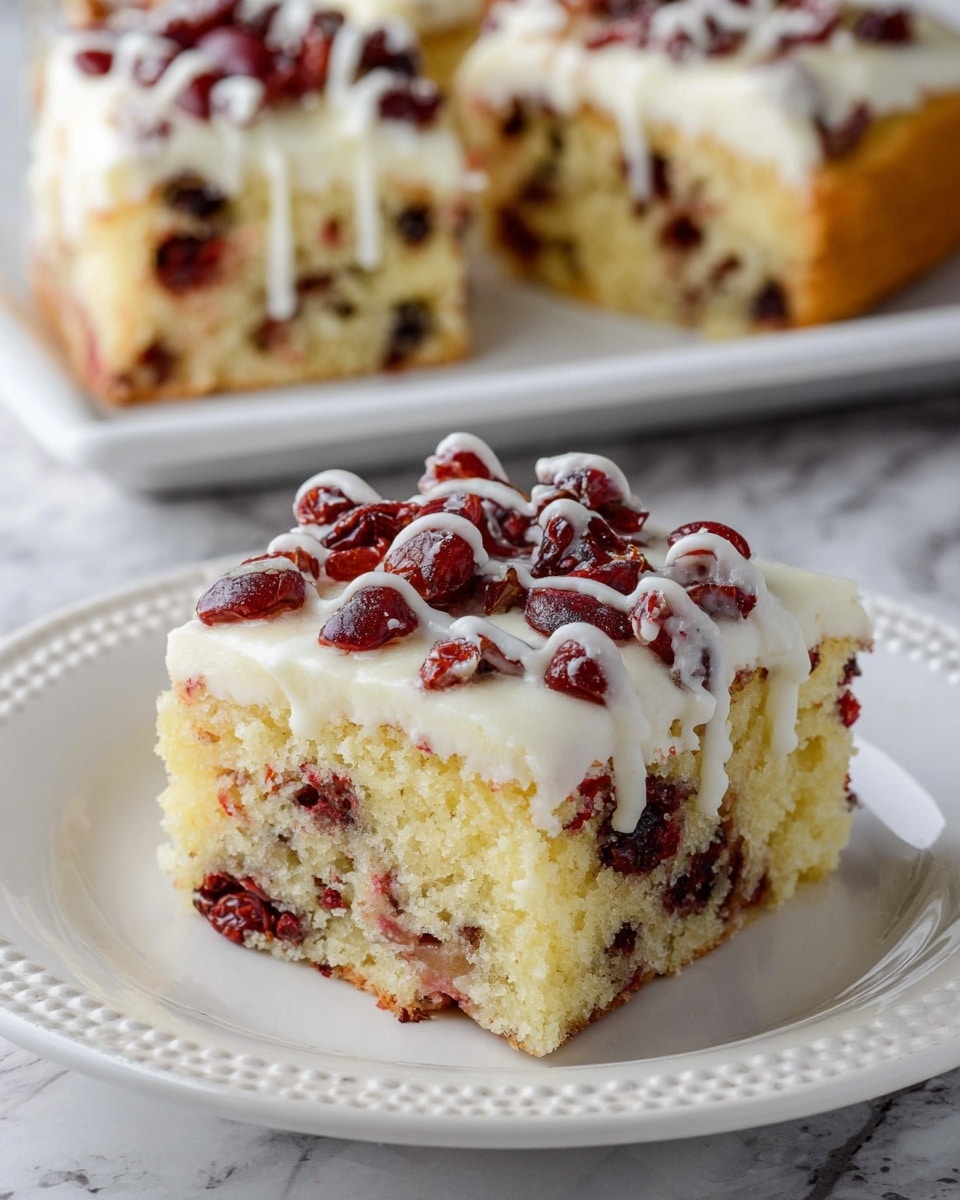 The image shows a square piece of cake on a white plate with a decorative edge, placed on a white marbled surface. The cake has two visible layers; the bottom layer is light yellow with red dried fruit pieces mixed inside, while the top layer is a smooth white frosting, covered with whole red dried fruit pieces and drizzled with thin white icing. In the background, there are two more pieces of the same cake, slightly blurred, on a white rectangular plate. Photo taken with an iphone --ar 4:5 --v 7