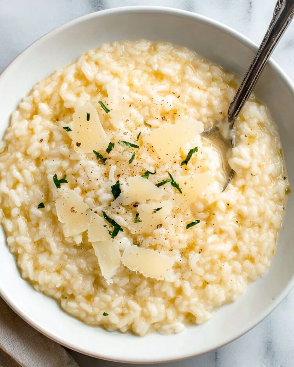 A close-up of a creamy risotto served in a white bowl, featuring a smooth, pale beige layer of rice with a slightly glossy texture. The risotto is topped with thin, off-white cheese shavings scattered across the center. Sprinkled finely chopped green herbs add a light contrast on top. A metal spoon rests inside the bowl on the right side, partially submerged in the risotto. The bowl sits on a white marbled surface. photo taken with an iphone --ar 4:5 --v 7