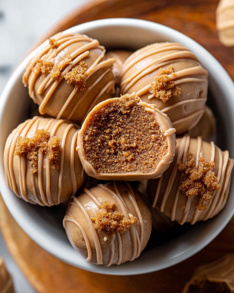 A close-up view of a white bowl filled with several round truffles coated in a smooth, light caramel-colored shell with delicate zigzag drizzle lines of the same caramel color on top. One truffle is cut in half and held above the others, revealing a soft, crumbly, textured brown filling inside. Small crumbs of the filling are sprinkled lightly on top of some truffles. The bowl is placed on a wooden tray, all set against a white marbled surface. Photo taken with an iphone --ar 4:5 --v 7