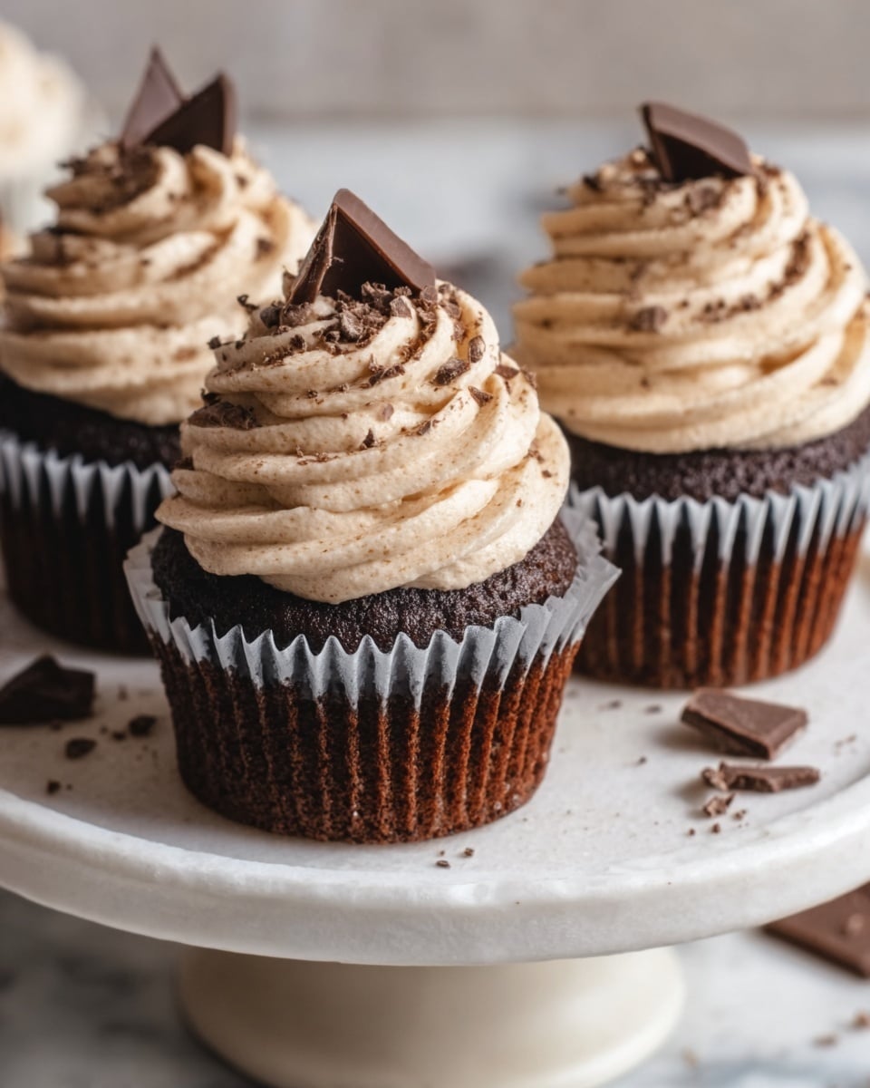 Three chocolate cupcakes sit on a white cake stand with a white marbled surface below. Each cupcake has a dark brown base with a swirl of light beige cream topping, piped in tall spirals. On top of each swirl is a small roasted coffee bean, and around the base are tiny chocolate flakes scattered on the cake stand. In the background, the white marbled texture is softly blurred. Photo taken with an iphone --ar 4:5 --v 7