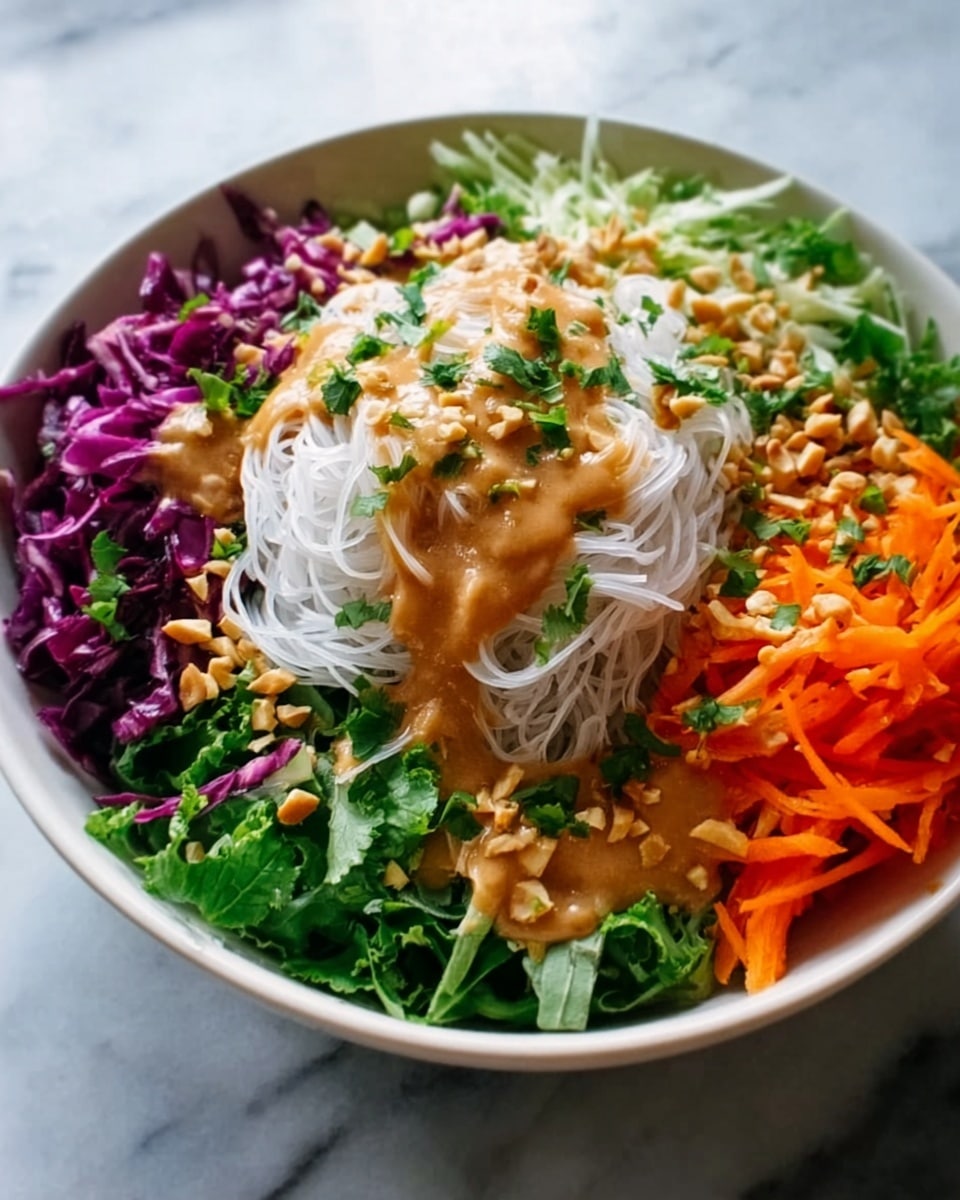 A white bowl filled with a vibrant layered salad set on a white marbled surface. The bottom layer consists of shredded green leafy vegetables, topped with a neat pile of white rice noodles in the center. To the left of the noodles, there is a bright purple shredded cabbage, and on the upper right side, thin orange carrot strips are arranged. The entire bowl is drizzled generously with a smooth, light brown creamy sauce, sprinkled with chopped green herbs and crushed nuts. The colors are fresh and bright, and the textures range from crispy vegetables to soft noodles and creamy sauce. Photo taken with an iphone --ar 4:5 --v 7