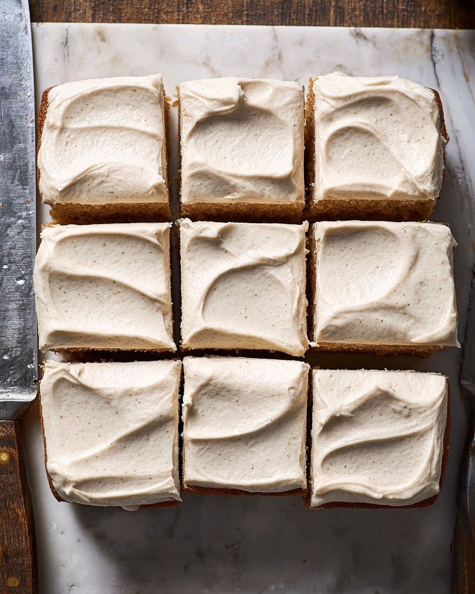 The image shows a square cake cut into nine pieces, arranged in a 3 by 3 grid. Each piece has one layer of light brown cake at the bottom and a thick, even layer of creamy, light beige frosting spread smoothly on top. The frosting has soft swirls and a slightly textured appearance with small specks, giving it a natural look. The cake sits directly on a white marbled surface, with a slight shadow around the edges. Part of a metal baking tray and a knife are visible on the bottom left side of the image. photo taken with an iphone --ar 4:5 --v 7