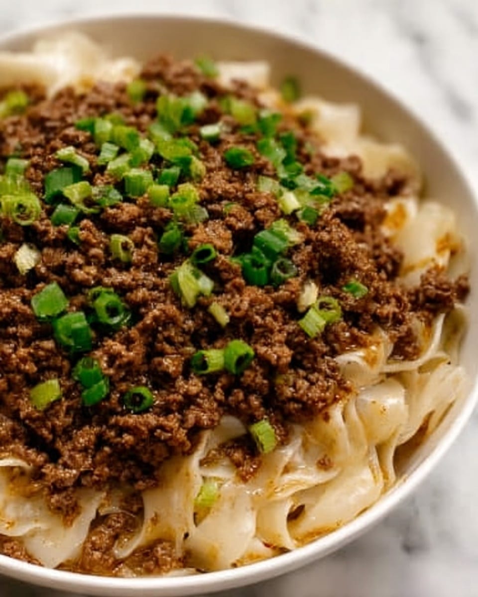 The image shows a close-up of a bowl filled with cooked wide noodles that look soft and slightly shiny, forming the base layer. On top of the noodles, there is a thick layer of crumbled cooked ground beef that is brown and textured. Scattered evenly over the beef are small pieces of chopped green onions, adding bright green color to the dish. The bowl is white and sits on a white marbled surface. photo taken with an iphone --ar 4:5 --v 7