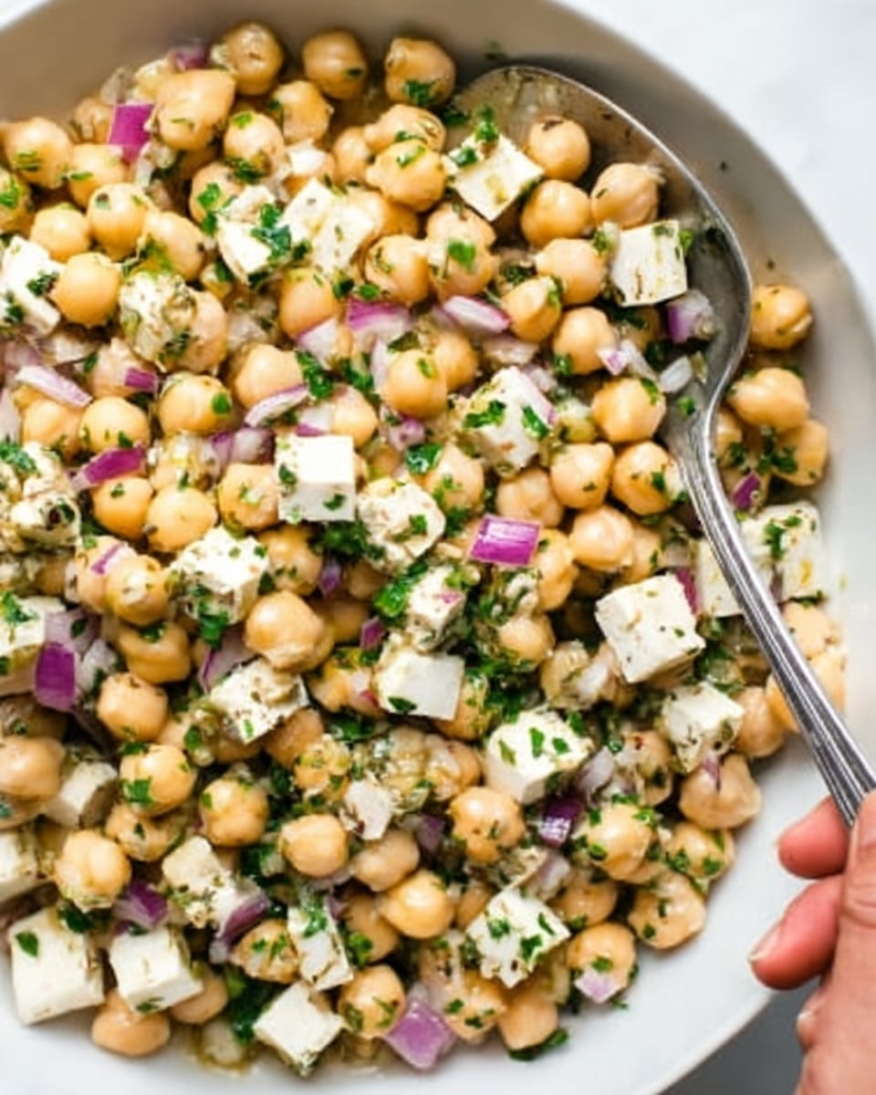 The image shows a close-up of a white bowl filled with a colorful chickpea salad. The salad has three main layers: the light beige chickpeas that cover most of the bowl, white cubes of cheese scattered evenly, and small pieces of purple onion mixed throughout. Bright green chopped herbs are sprinkled all over the salad, adding freshness and color contrast. A silver spoon is placed inside the bowl on the right side, with a woman's hand holding it, ready to serve. The background is a white marbled surface that adds a clean and bright look to the image. Photo taken with an iphone --ar 4:5 --v 7