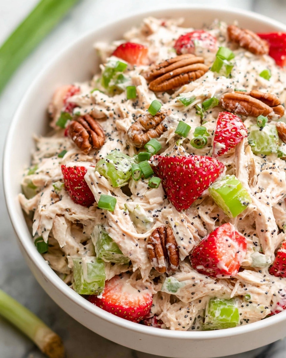 A close-up view of a white bowl filled with a creamy chicken salad, showing several layers and textures: shredded pale beige chicken mixed with light cream sauce forms the main layer, dotted with bright red strawberry chunks, bright green celery pieces, and dark brown pecans; vibrant green chopped spring onions and tiny black poppy seeds are sprinkled throughout, adding color contrast and texture. The bowl sits on a white marbled surface, emphasizing the colors of the salad. photo taken with an iphone --ar 4:5 --v 7