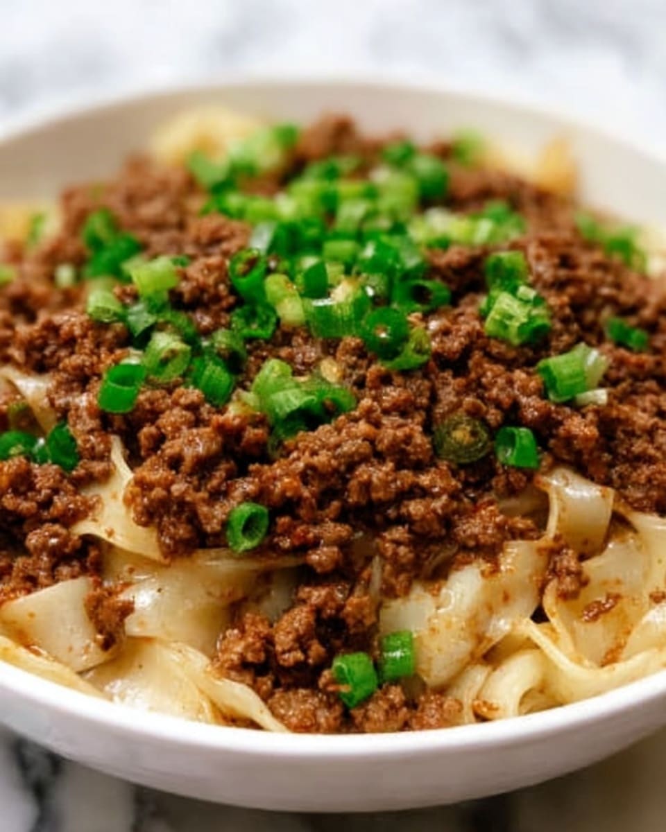 A white bowl filled with a layered dish showing soft, wide noodles at the bottom, slightly yellow and shiny from sauce. On top, there is a thick layer of cooked ground meat, rich brown with a crumbly texture. Scattered over the meat are small white bits, likely onions, mixed with chopped green onions that add fresh green color. The dish looks warm and hearty with a mix of smooth noodles and coarse meat pieces, all set against a white marbled surface. Photo taken with an iphone --ar 4:5 --v 7