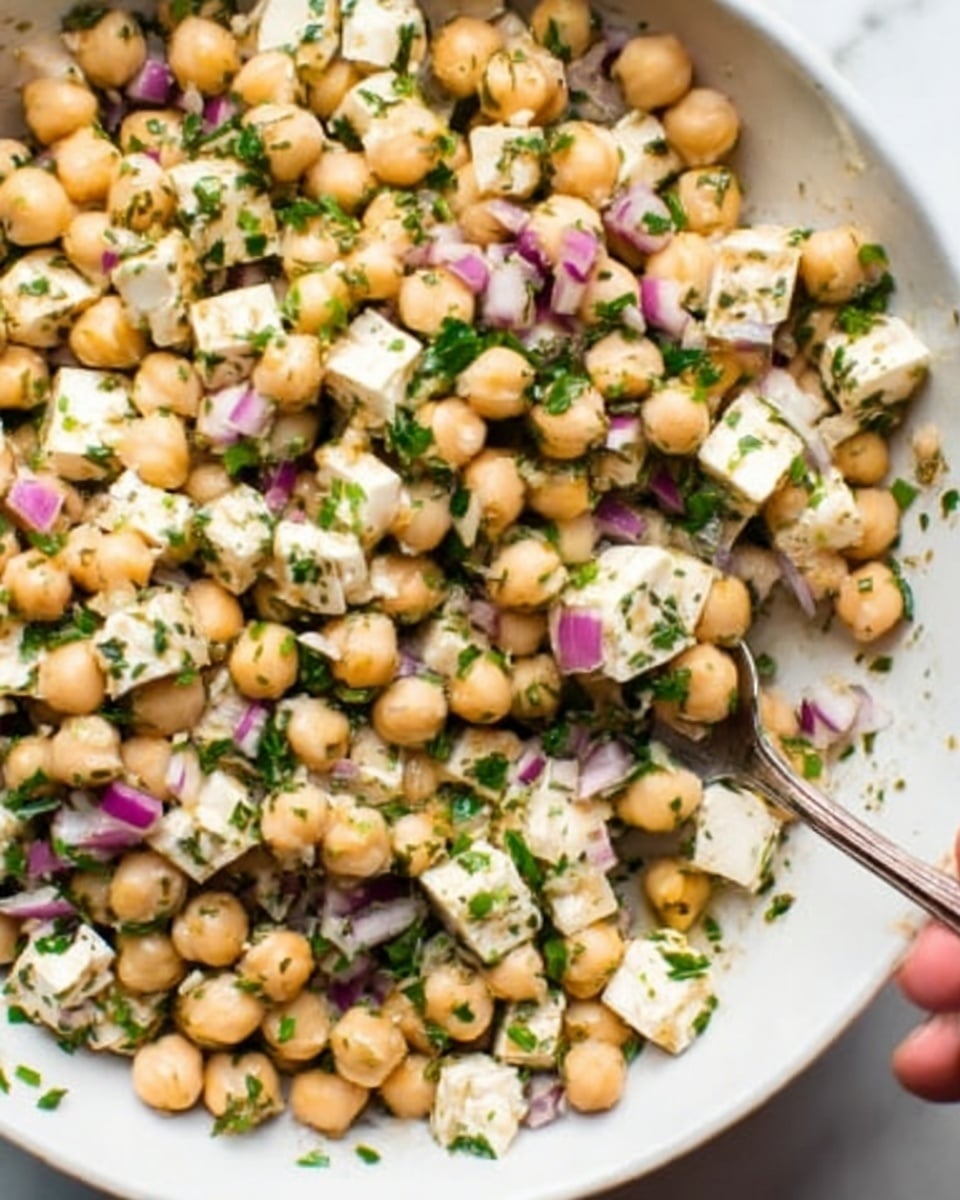 The image shows a close-up of a white bowl filled with a chickpea salad. The salad has three main layers: light beige chickpeas, small white cubes of cheese, and bits of purple-red onion mixed with green chopped herbs. The chickpeas are round and smooth, the cheese cubes are firm with a smooth surface, and the onion pieces add a touch of crispness and color contrast. All ingredients are evenly mixed, giving a fresh and colorful look. A silver spoon is partially visible inside the bowl, with a woman's hand holding it at the edge. The background is a white marbled surface. photo taken with an iphone --ar 4:5 --v 7