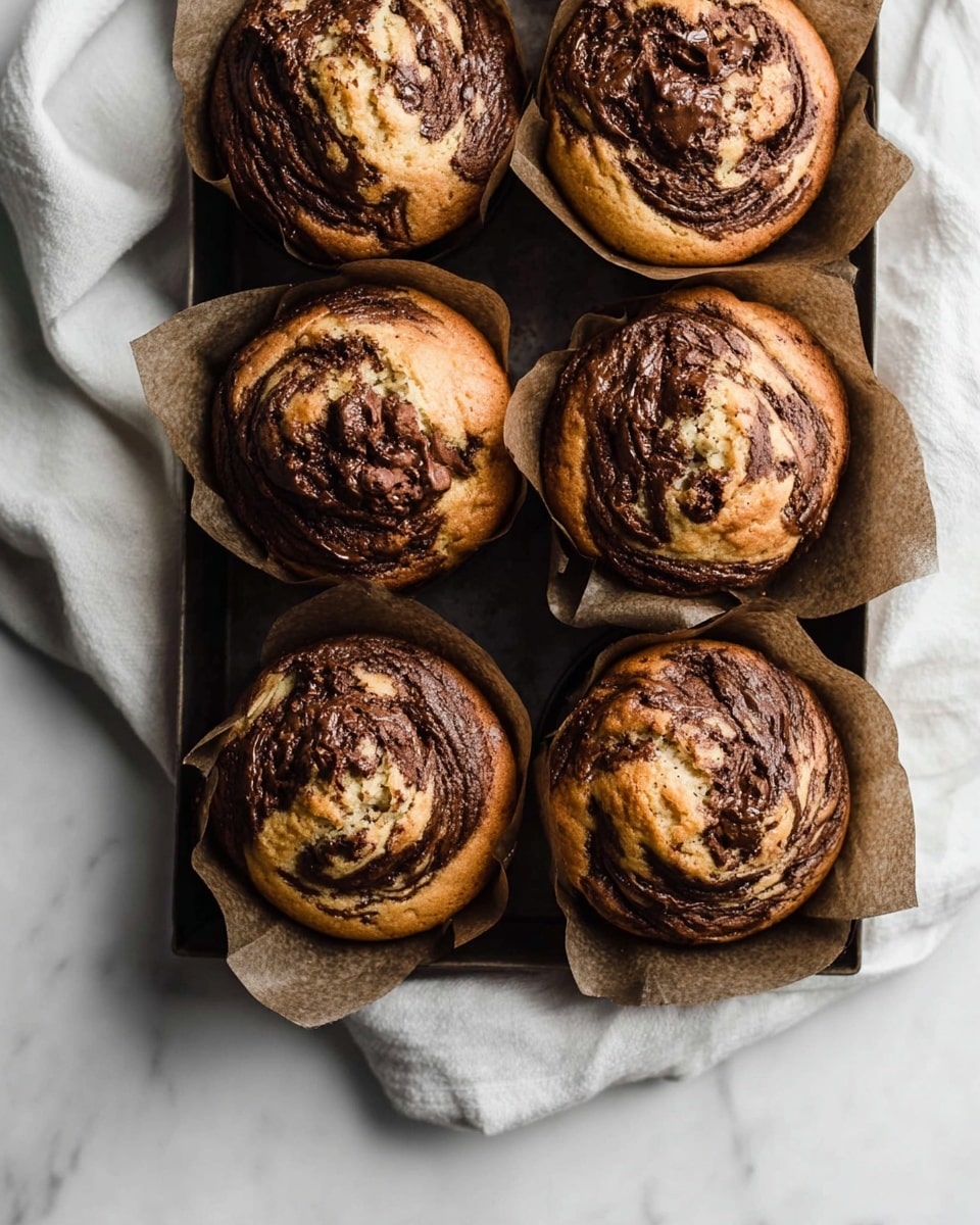 Six muffins with golden brown tops and swirls of dark chocolate are arranged in two rows of three inside dark brown paper wrappers. The muffins have a soft, slightly crinkled texture where the chocolate is swirled in, creating a marbled pattern on each one. They sit on a soft, light gray cloth that is gently crumpled, all placed on a white marbled surface. The light is natural and soft, highlighting the warm tones of the muffins and the smooth texture of the chocolate. photo taken with an iphone --ar 4:5 --v 7