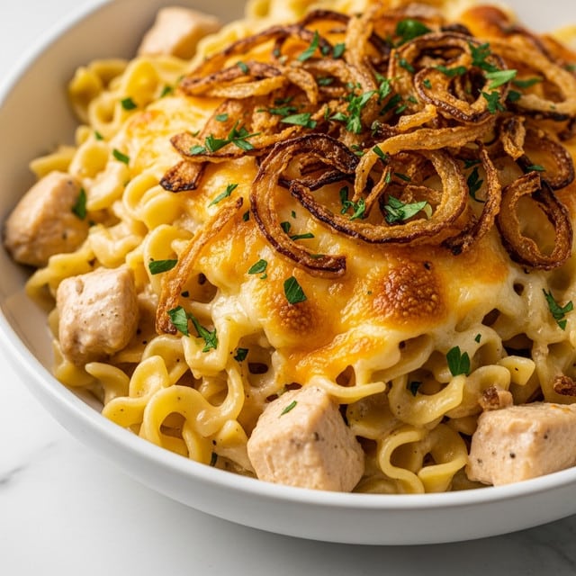 A close-up view of a creamy noodle dish served in a white bowl placed on a white marbled surface. The dish has multiple layers starting with a base of soft, yellow noodles mixed with chunks of cooked chicken. On top of the noodles, there is a layer of melted cheese that appears golden and slightly bubbly. The surface is topped with crispy, browned fried onions that add a crunchy texture, along with sprinkled small green parsley leaves for a fresh touch. The lighting highlights the rich creaminess and golden brown crispy details, making the dish look warm and inviting. Photo taken with an iphone --ar 4:5 --v 7