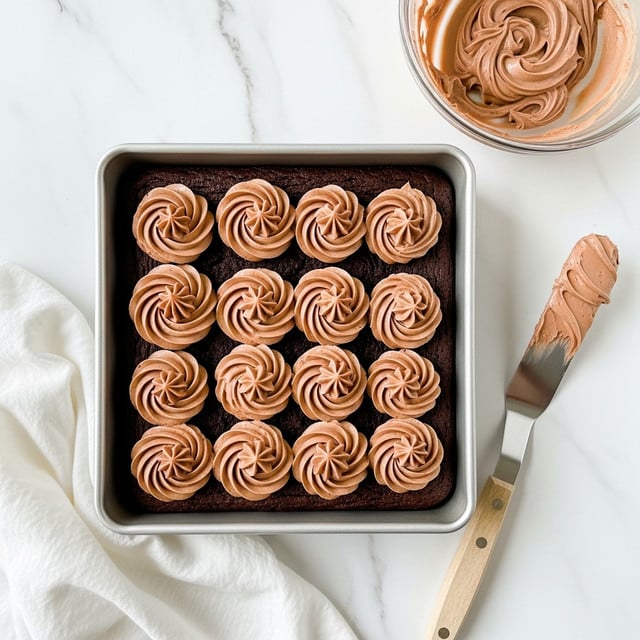 A square chocolate brownie sits inside a metal baking pan with a thick layer of creamy chocolate frosting spread on top in large round dollops, creating an uneven textured surface with swirls and peaks. Next to the pan is a glass bowl filled with smooth chocolate frosting, showing a swirl pattern, and a spatula with a wooden handle covered in some frosting lying flat on the white marbled surface. A white cloth is partially tucked underneath the pan at the bottom left corner. Photo taken with an iphone --ar 4:5 --v 7