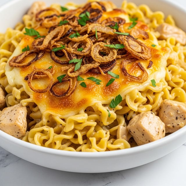 A close-up view of a white bowl filled with creamy baked noodles mixed with shredded chicken, creating a soft, light beige base layer. On top of this is a golden melted cheese layer that adds a smooth and slightly crispy texture. The dish is topped with thin, crispy fried onions in shades of deep golden brown, scattered unevenly over the entire surface. Small green parsley bits are sprinkled on top, adding a fresh pop of color. The bowl sits on a white marbled surface, and the photo is taken with an iphone --ar 4:5 --v 7