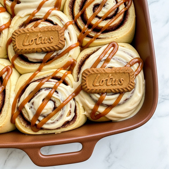 The image shows four cinnamon rolls placed closely in a brown baking dish against a white marbled textured background. Each roll has a soft golden-brown dough base with visible swirls of darker cinnamon filling. The rolls are covered in a thick, creamy beige icing that looks smooth and glossy, with a rich caramel drizzle artistically zigzagged on top adding a deeper brown color contrast. Three of the rolls have a small, rectangular brown biscuit with a textured pattern sitting on top. The dish handle is visible at the bottom of the image, and the focus is close up, showing the gooey texture of the icing. photo taken with an iphone --ar 4:5 --v 7