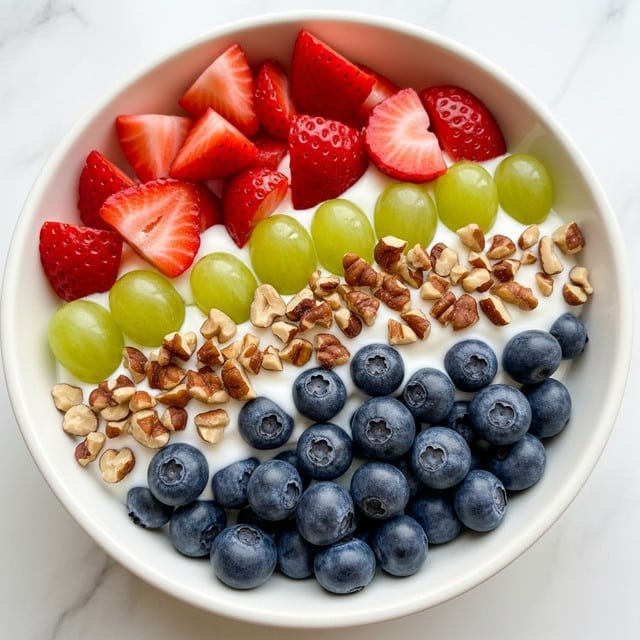 A white bowl filled with a fresh fruit salad made of three main layers: large chunks of red strawberries and light green grapes, small round blueberries, and white cream that coats the fruit pieces giving them a smooth, shiny look. On top of these fruit layers are scattered medium-sized chopped brown nuts, adding a crunchy texture. The bowl rests on a white marbled surface, and the salad looks colorful and fresh with a mix of bright red, green, blue, and creamy white colors photo taken with an iphone --ar 4:5 --v 7