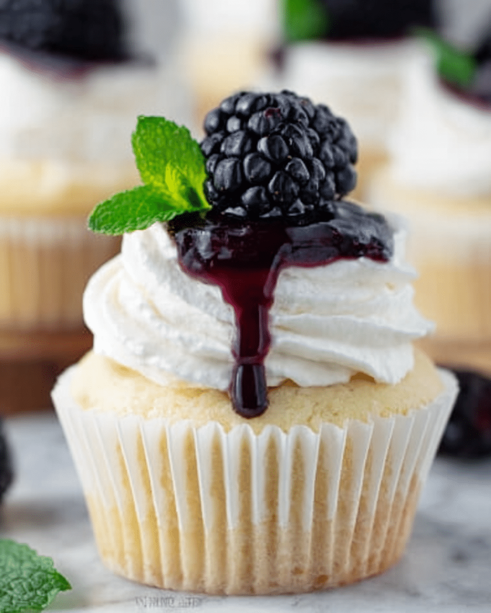 A close-up of a single cupcake in a white paper liner on a white marbled surface shows three clear layers. The bottom layer is a light yellow vanilla cake with a soft and moist texture. On top of the cake is a thick layer of white whipped cream, smooth and fluffy, piped in a swirl. Crowning the cupcake is a glossy black blackberry with deep purple juice dripping down the whipped cream, accompanied by a small bright green mint leaf for contrast. The background slightly blurs other similar cupcakes. Photo taken with an iphone --ar 4:5 --v 7