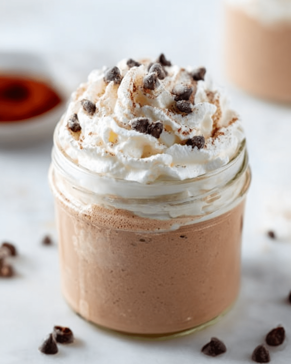 The image shows a glass jar filled with three visible layers. The bottom layer is a light brown creamy mousse with a smooth texture. Above this mousse is a swirl of white whipped cream with soft peaks. On top of the whipped cream are small dark chocolate chips and light brown coffee powder scattered around for decoration. The jar sits on a white marbled surface, and a blurred light-colored background is visible. photo taken with an iphone --ar 4:5 --v 7
