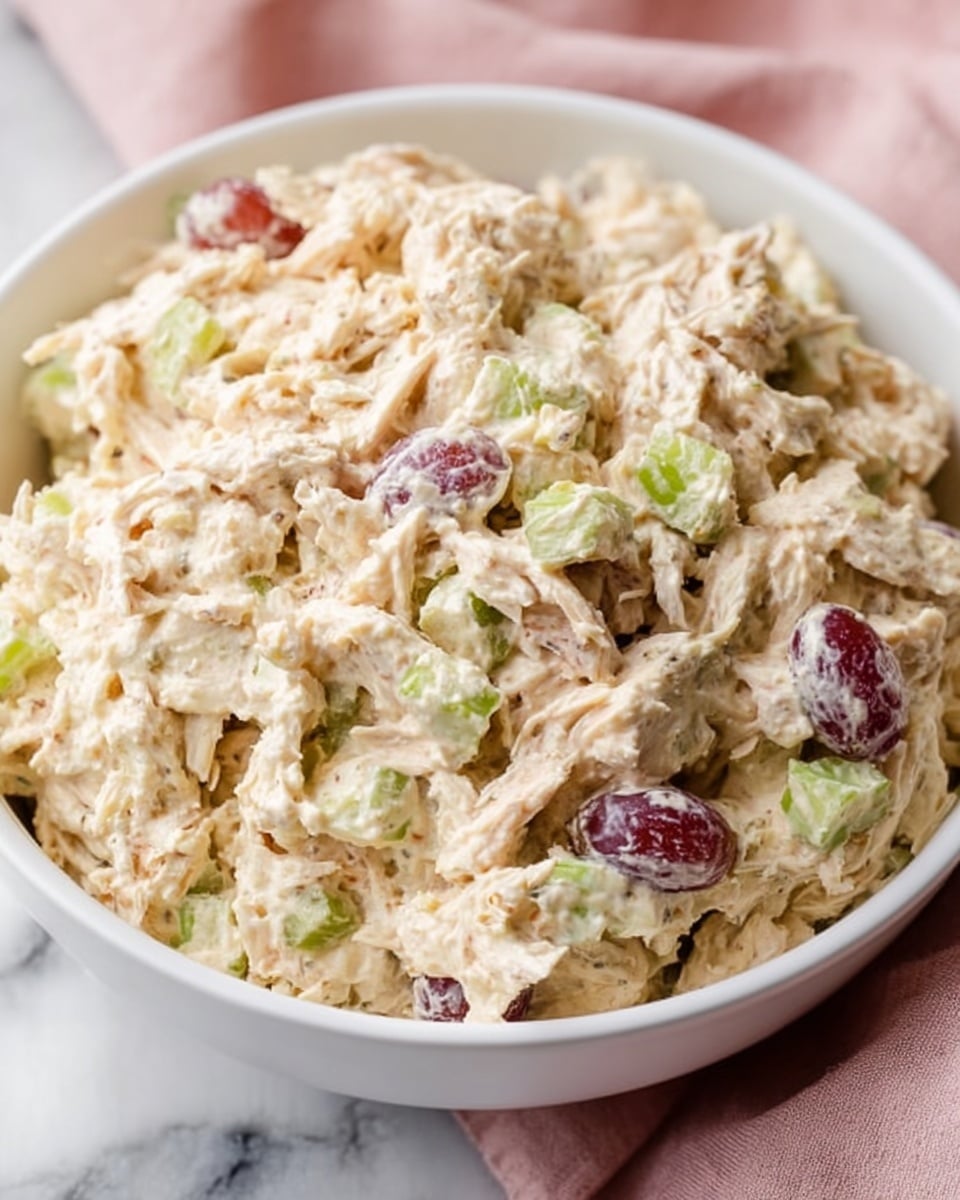 A close-up of a white bowl filled with a creamy shredded chicken salad, showing a mixture of light beige shredded chicken, small chunks of green celery, and pieces of red grapes scattered throughout. The texture looks soft and moist, with the ingredients fully mixed but still visible separately. The bowl is placed on a soft pink cloth that sits on a white marbled surface, creating a gentle and cozy background. The whole image has a warm and inviting feel. Photo taken with an iphone --ar 4:5 --v 7