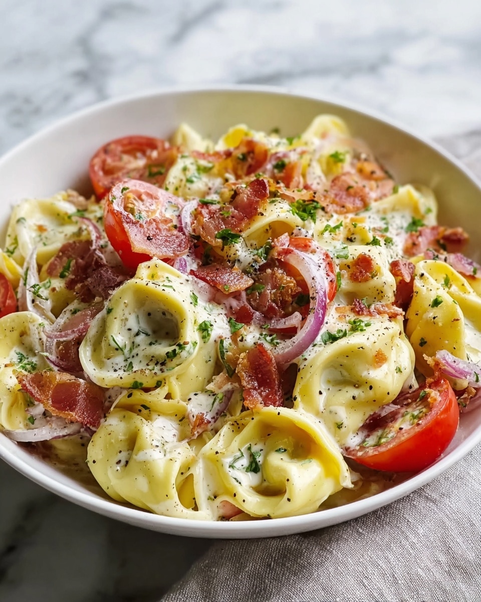 The image shows a white bowl filled with creamy tortellini pasta, each piece pale yellow with a smooth, soft texture, curled and stuffed. Mixed in are vibrant red cherry tomato halves adding bright color, along with crispy pieces of bacon that have a rich golden-brown and slightly crunchy look. Thin slices of light purple onion are scattered throughout, adding an extra layer of texture. The dish is sprinkled with finely chopped green herbs and cracked black pepper, all sitting on a white marbled surface. photo taken with an iphone --ar 4:5 --v 7