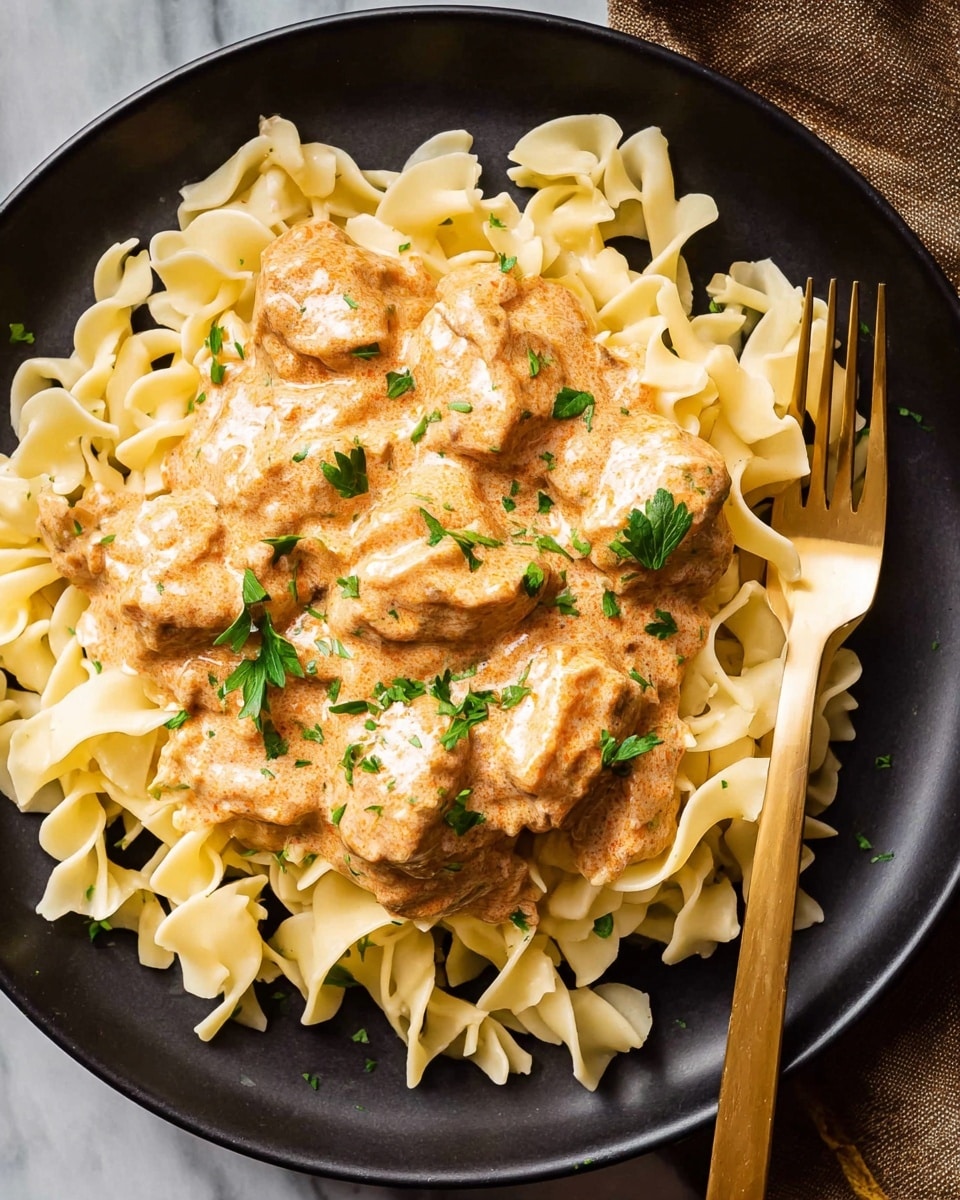 A dish featuring light yellow egg noodles forming a loose, circular base layer on a white plate, topped with medium-sized, irregularly shaped pieces of chicken in a rich, creamy orange-brown sauce that lightly covers the noodles around the edges. The sauce has a smooth texture with some visible specks of seasoning. Small, fresh green parsley leaves are scattered evenly across the chicken and sauce, adding a pop of color. To the left, a gold-colored fork rests on the edge of the plate. The entire setup sits on a white marbled textured surface. photo taken with an iphone --ar 4:5 --v 7