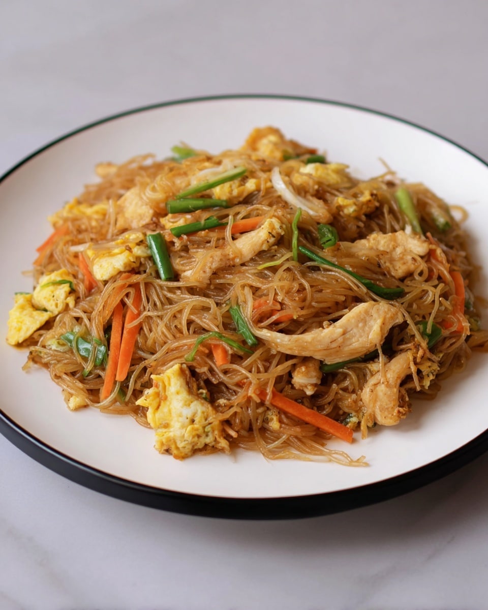 A white plate with a black rim holds a serving of stir-fried noodles made of thin, lightly browned strands mixed with thin green and orange vegetable strips, such as bell peppers and carrots. There are several pieces of cooked chicken that are light golden-brown on top of and mixed throughout the noodles. Small, irregular pieces of scrambled egg with a soft texture and light yellow color are scattered evenly in the dish. The plate sits on a white marbled surface with a soft, natural light shining on the food, giving it a warm, fresh look. photo taken with an iphone --ar 4:5 --v 7