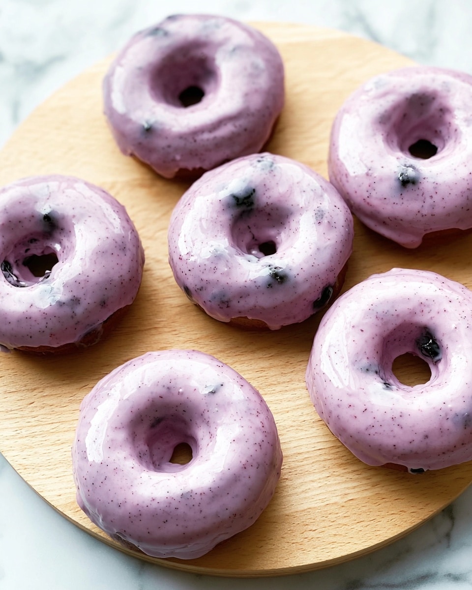The image shows five round, golden-brown doughnuts dusted with white powdered sugar arranged on a dark board, set against a white marbled surface. Each doughnut has a hollowed-out center filled with bright red strawberry jam, topped with a whole, glossy, fresh strawberry with green leaves. One doughnut in the front is cut open, revealing a creamy white filling with bits of strawberry inside. Around the doughnuts, there are fresh strawberries and small green leaves scattered for decoration. The soft lighting enhances the warm colors and the moist texture of the doughnuts and strawberries. Photo taken with an iphone --ar 4:5 --v 7