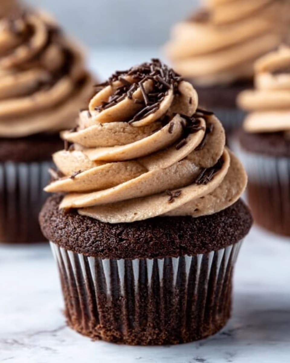 A chocolate cupcake with a dark brown base topped with a tall swirl of light brown chocolate frosting, sprinkled with small dark chocolate shavings on top; the cupcake is wrapped in a dark brown paper liner, sitting on a white marbled surface with similar cupcakes blurred in the background. photo taken with an iphone --ar 4:5 --v 7