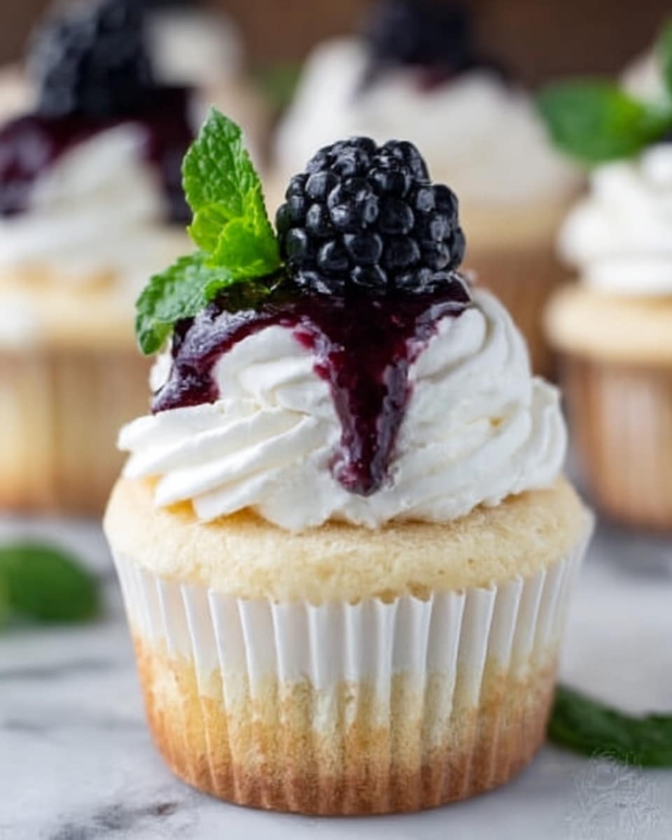 A small vanilla cupcake in a white paper liner sits on a white marbled surface. The cupcake has two layers of creamy white frosting, the bottom layer covering the cupcake top smoothly and the second layer piped thick and swirled above it. On top of the frosting sits a fresh black blackberry with a small bright green mint leaf behind it. Dark red berry sauce drips slightly down the side of the frosting. The background is softly blurred showing more similar cupcakes. Photo taken with an iphone --ar 4:5 --v 7