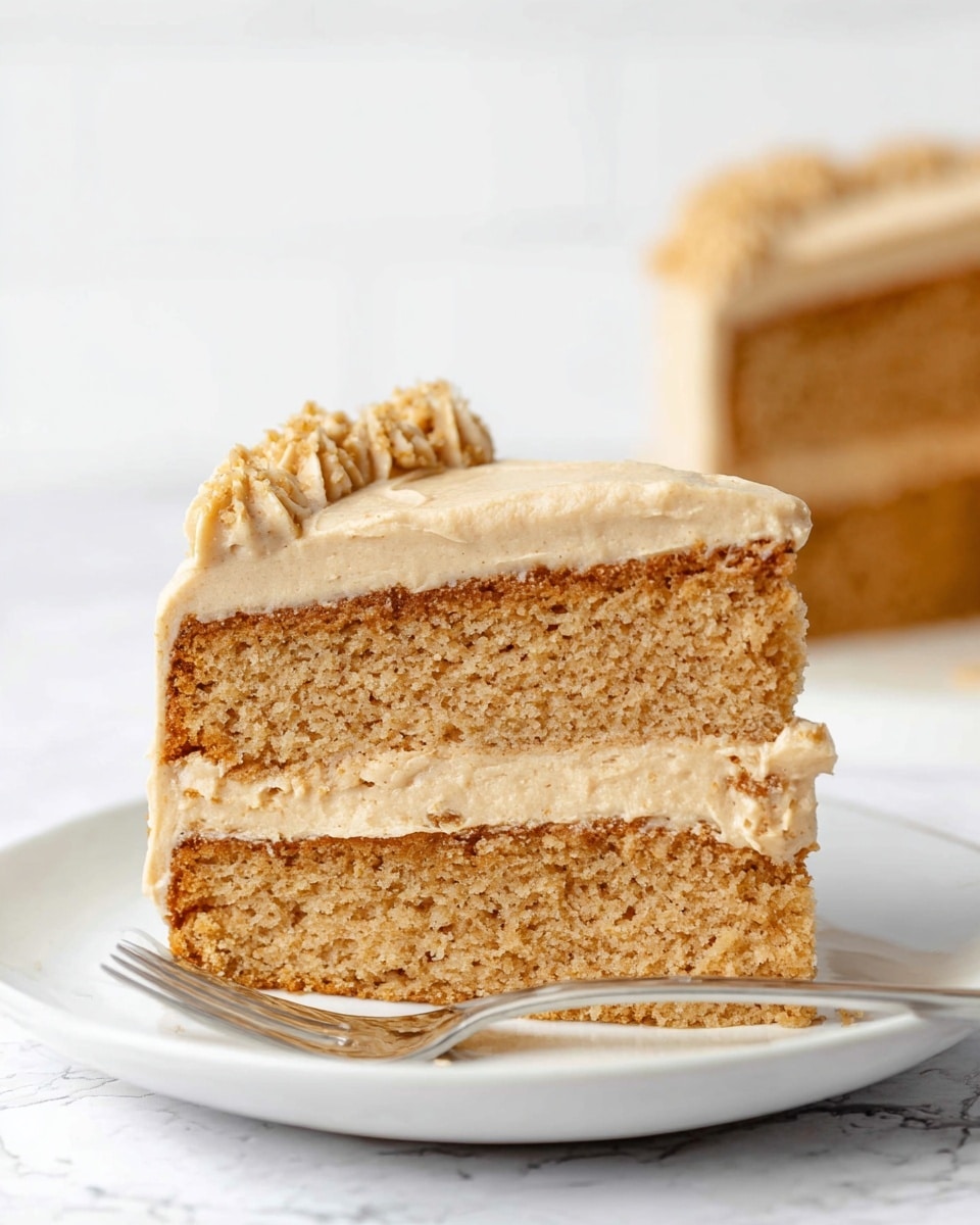 The image shows a slice of two-layer cake on a white plate with a white marbled background. The cake layers are light brown and moist in texture. Between the two layers, there is a thicker layer of creamy frosting that is a smooth light tan color, and on top, there is another layer of the same frosting, neatly spread with some textured crumbs or small decoration pieces on the edge. The cake looks soft and well-risen. A silver fork is placed in front of the plate. Photo taken with an iphone --ar 4:5 --v 7