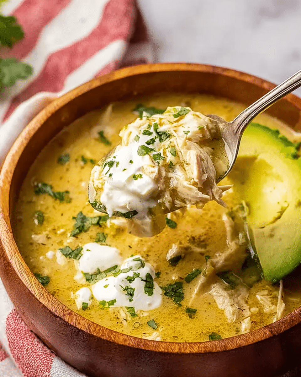 A wooden bowl filled with thick yellow soup mixed with shredded white chicken pieces and green herbs mixed in. The soup surface has dollops of white cream and some green cilantro sprinkled on top. A slice of avocado with a smooth green flesh rests on the right side inside the bowl. A spoon lifting a scoop of soup shows creamy texture with white chicken, green herbs, and cream. The background has a blurred white marbled texture with a red and white kitchen towel visible. Photo taken with an iphone --ar 4:5 --v 7