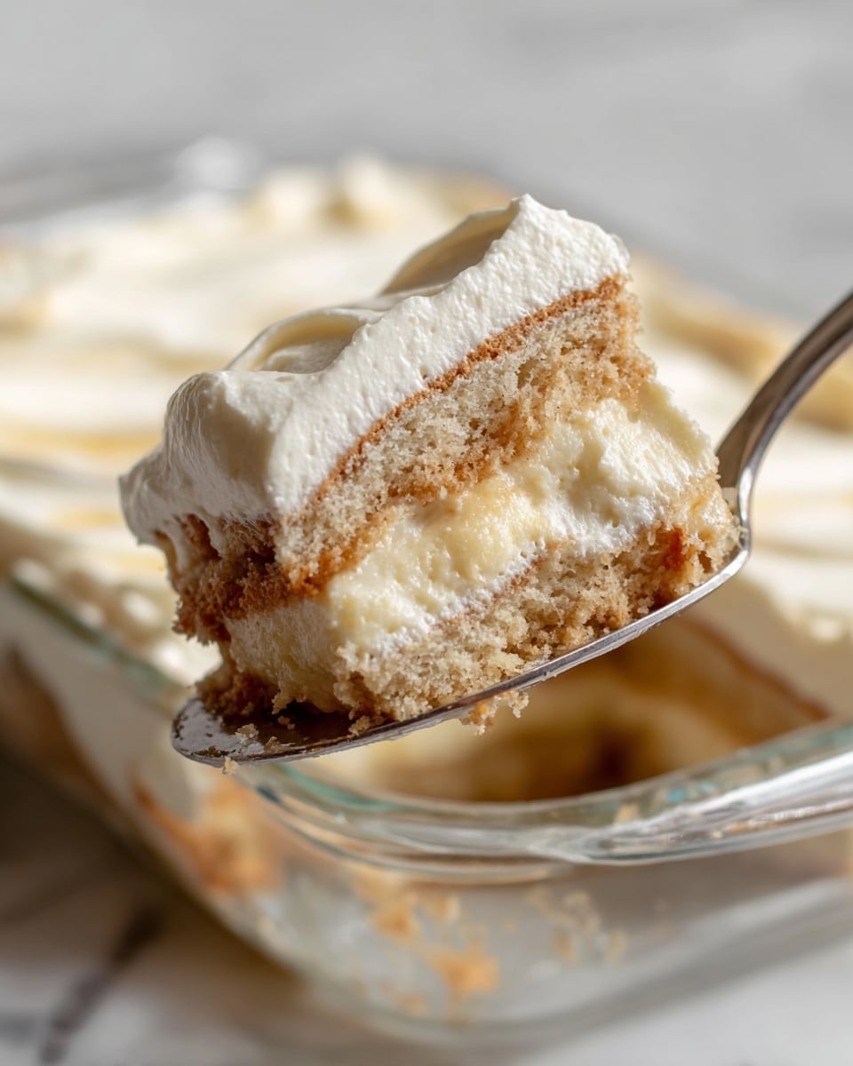 A close-up view of a three-layer slice of creamy cake held by a silver spoon inside a clear glass dish. Each layer of the cake has a light beige, soft and spongy texture with smooth, thick white cream filling between layers and covering the top. The cake shows sponge edges slightly soaked, giving a moist look, with a fluffy and airy cream that appears rich and smooth. The background has a white marbled texture that adds a clean and bright feel to the image. Photo taken with an iphone --ar 4:5 --v 7