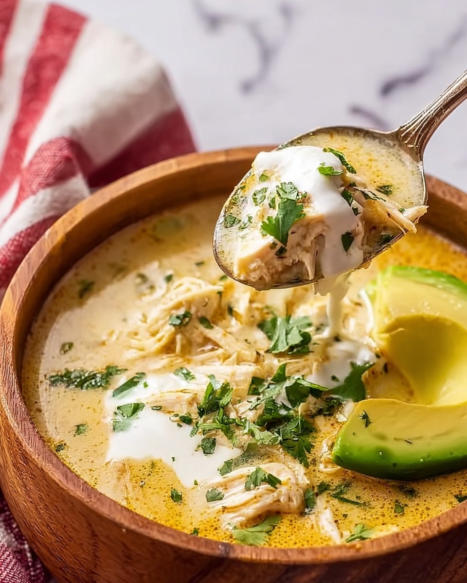 A close-up view of a white bowl filled with creamy chicken soup showing several layers: a smooth light yellow creamy broth with visible green herb bits throughout, tender white shredded chicken mixed into the soup, dollops of white sour cream on top, and a fresh bright green avocado slice resting on the side of the bowl. A silver spoon held by a woman's hand is scooping up a portion of the soup with chicken, sour cream, and herbs. The bowl is placed on a white marbled surface with a soft red and white cloth partially visible in the background. Photo taken with an iphone --ar 4:5 --v 7
