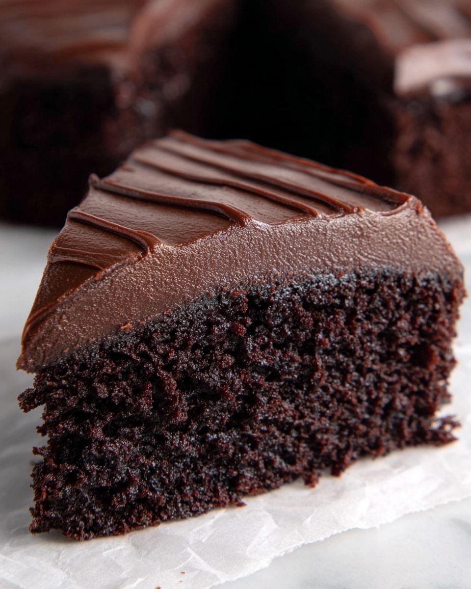 A close-up of a single slice of rich chocolate cake sits on white parchment paper over a white marbled surface. The cake has two layers: the bottom layer is dark brown, moist, and crumbly with a spongy texture, while the top layer is thick, smooth chocolate frosting with a shiny finish and gentle diagonal lines etched across it, creating a subtle pattern. The background shows blurred parts of more chocolate cake pieces. Photo taken with an iphone --ar 4:5 --v 7