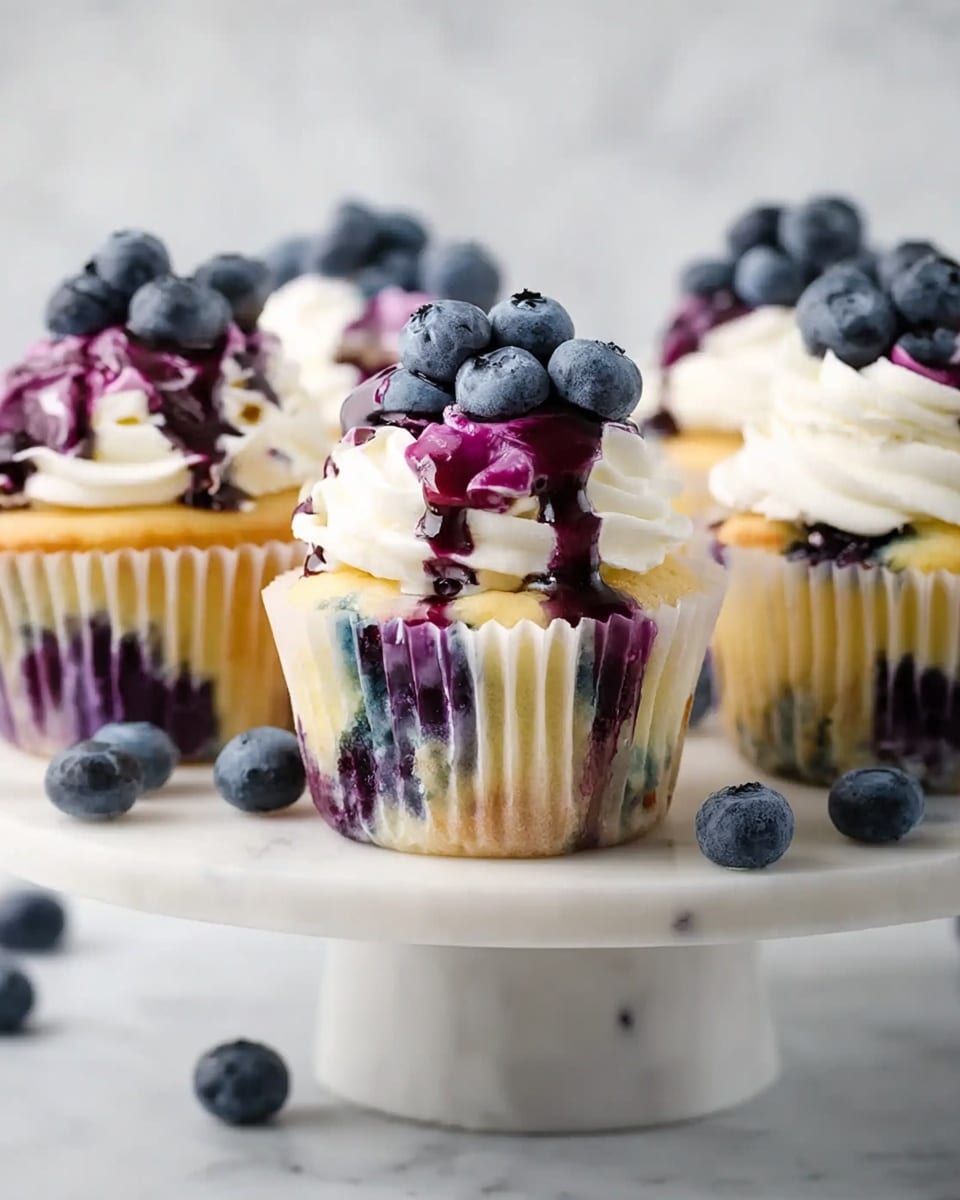 The image shows four yellow cupcakes with a slightly golden top, each wrapped in a white paper liner. Each cupcake has a swirl of white cream frosting on top, decorated with a drizzle of thick, dark purple blueberry sauce that runs down the sides. Fresh blueberries are placed on the frosting peak, adding a rich blue color contrast. The cupcakes are arranged on a round cake stand with a white marbled surface, with a few loose blueberries scattered around. The background is light and simple, highlighting the cupcakes clearly. Photo taken with an iphone --ar 4:5 --v 7