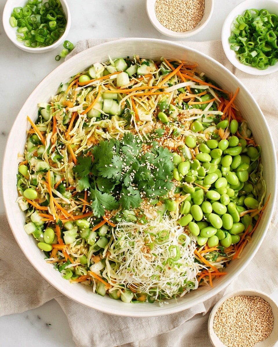 A large white bowl filled with a colorful fresh salad resting on a white marbled texture with a beige linen cloth underneath. The salad has a base layer of shredded light green cabbage, mixed with thin orange carrot sticks and bright green chopped cucumber pieces. On top, there is a vivid layer of whole green edamame beans and chopped green onions sprinkled throughout. Fresh green cilantro leaves are scattered generously, adding a leafy texture. The salad is finished with small white sesame seeds sprinkled evenly on top. Photo taken with an iphone --ar 4:5 --v 7