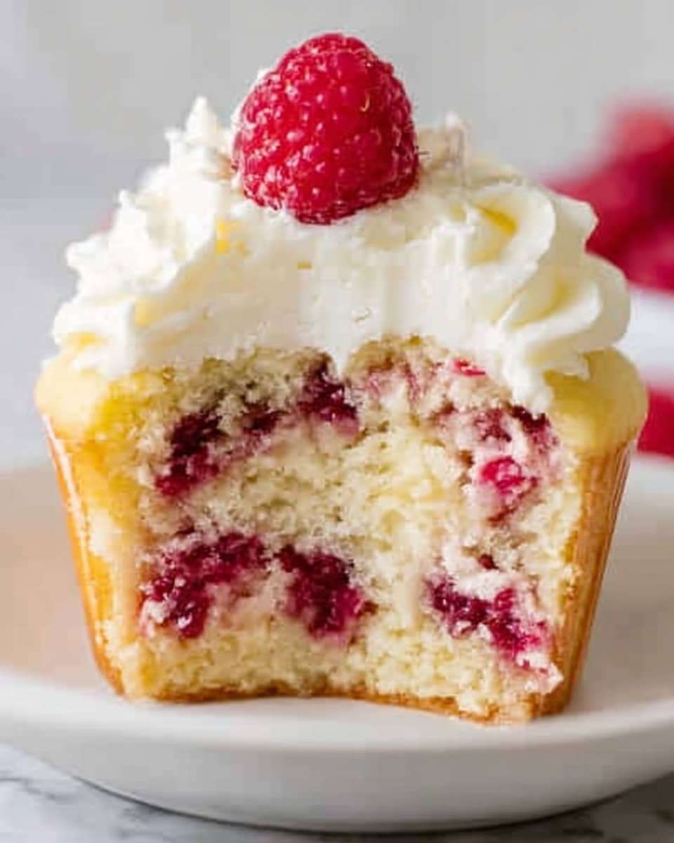 A close-up of a cupcake with three distinct layers is shown. The bottom layer is a pale yellow sponge cake with visible chunks of red raspberry inside, giving it a speckled texture. The middle layer contains creamy white frosting spread evenly, topped with another piece of light yellow sponge speckled with raspberries. The cupcake is finished with a generous swirl of white whipped frosting on top, crowned with a single bright red raspberry. The cupcake sits on a white plate, placed on a white marbled surface. photo taken with an iphone --ar 4:5 --v 7